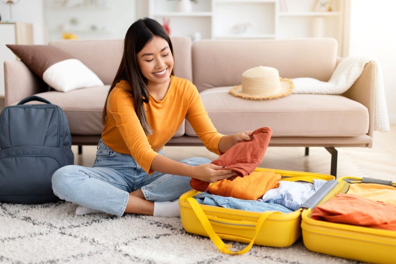 Cheerful young Chinese woman packing suitcase at home