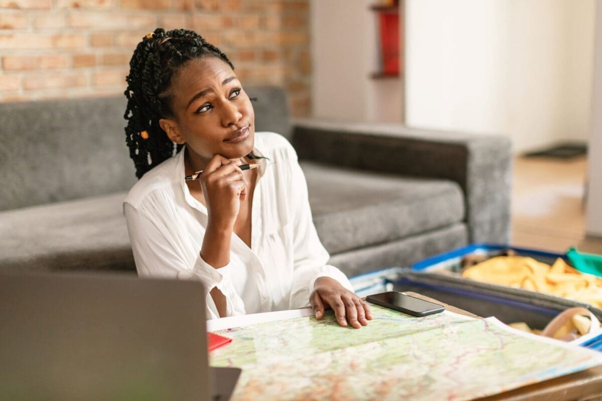 Dreamy African American woman planning future journey with map and laptop, thinking about best travel insurance essentials