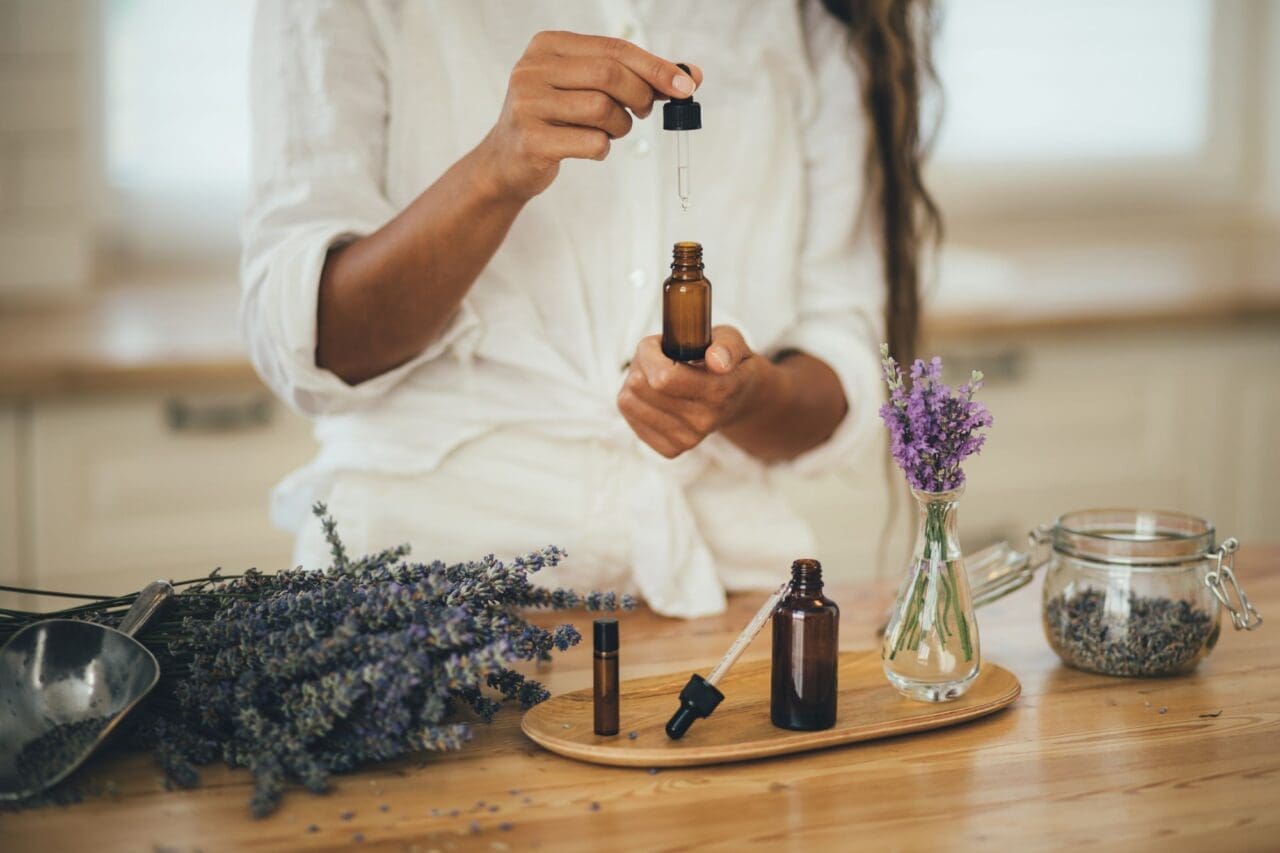 Woman holding an opened bottle of essential oil with lavender flowers and jars on the table